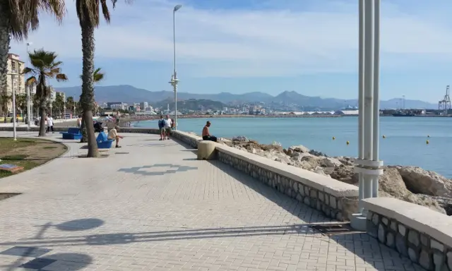 Ein Mann sitzt am Ufer der playa de la misericordia und blickt auf das Meer. Palmen säumen die Promenade, im Hintergrund die Stadt und Berge.