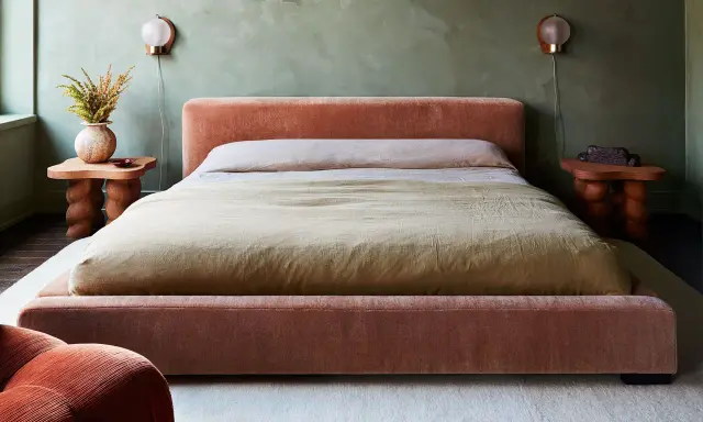 Cama king size tapizada en terciopelo rosa, con mesitas de noche de madera y lámparas de pared.