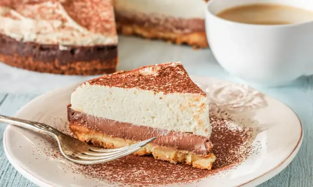 Pyszna tarta z mascarpone i czekoladą, posypana kakao, czeka na widelczyk. Obok filiżanka kawy.