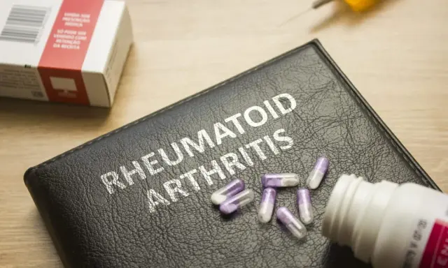 Na czarnej okładce z napisem "RHEUMATOID ARTHRITIS" leżą kapsułki. Obok znajduje się pudełko leków i strzykawka, sugerujące najnowsze leki na reumatoidalne zapalenie stawów.