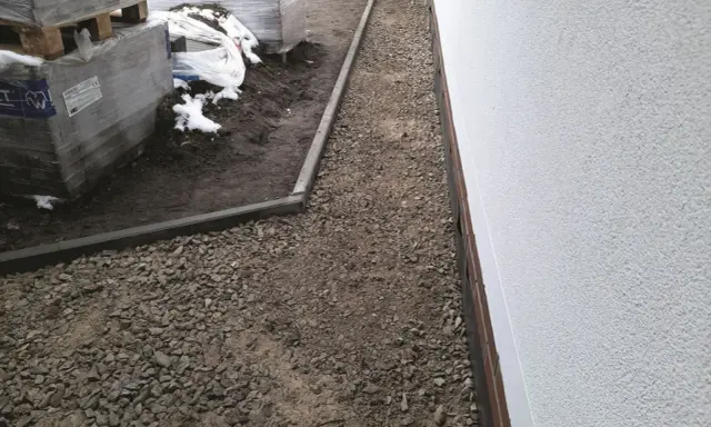 Budowa opaski wokół domu z kamienia i obrzeży. W tle palety z materiałami budowlanymi.
