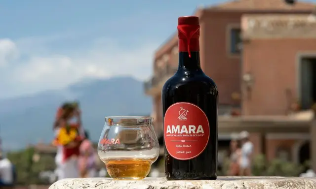 Bottiglia e bicchiere di Amara, un amaro che celebra come l'arancio amaro trama i sapori siciliani. Sfondo sfocato di un borgo.