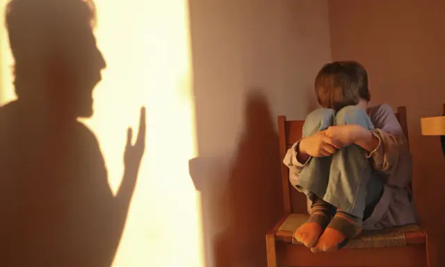 Cień ojca krzyczy, a dziecko skulone na krześle czuje się przytłoczone.