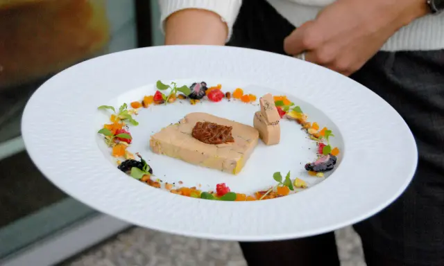 Une présentation raffinée de foie gras en entrée, ornée de fruits secs, de baies et de pousses vertes sur une assiette blanche texturée.