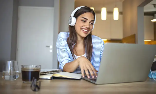 Uśmiechnięta studentka w słuchawkach pracuje przy laptopie. Czy muzyka pomaga w nauce? Ona chyba tak, bo wygląda na zrelaksowaną i skupioną.