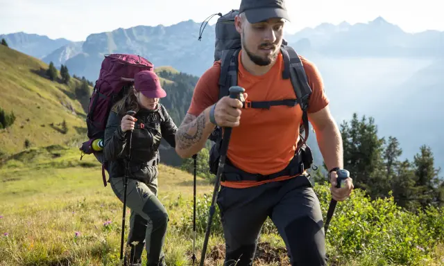 Dos excursionistas con mochilas y bastones caminan por una ladera montañosa.