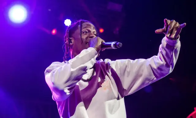 Travis Scott na scenie, w białej bluzie, z mikrofonem w dłoni. Jego energia inspiruje wielu **współautorów piosenek**.