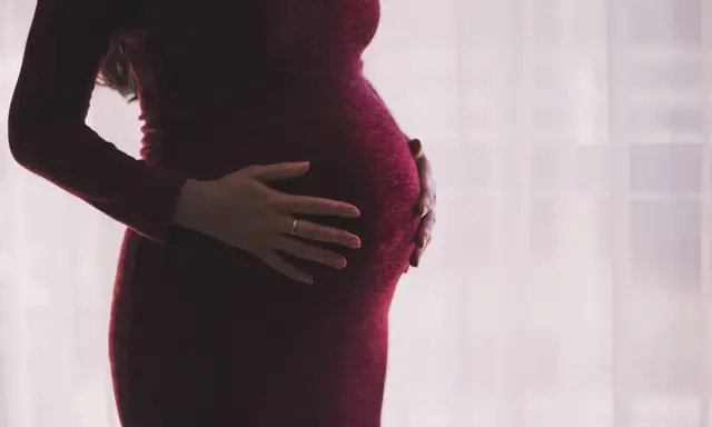 Kobieta w ciąży w bordowej sukience delikatnie obejmuje dłońmi brzuszek.