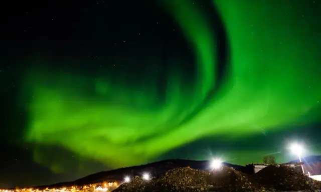 Spektakularna zorza polarna rozświetla nocne niebo nad ośnieżonymi wzgórzami i światłami miasteczka. To idealny moment, kiedy występuje zorza polarna.