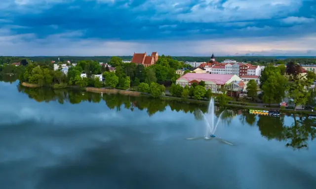 Widok na jezioro z fontanną i zabudowaniami, w tym kościołem. Odkryj iława atrakcje, które zachwycają o każdej porze dnia.