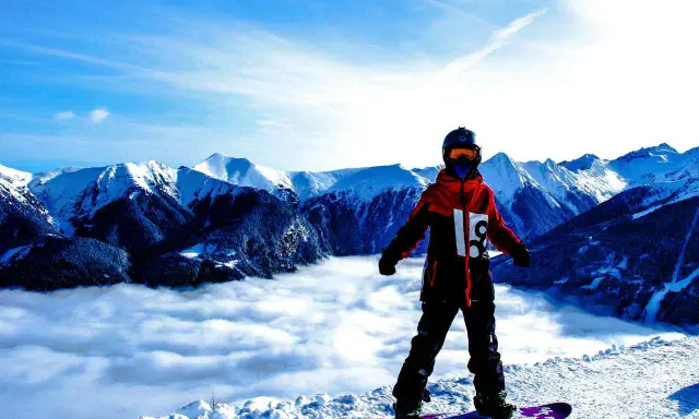 Snowboardzista w kurtce hardshell stoi na szczycie góry, nad morzem chmur. Góry w tle.