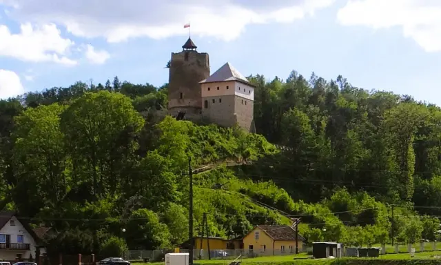 Zamek w Czchowie góruje nad zielonym wzgórzem, otoczony drzewami. Na szczycie wieży powiewa flaga.