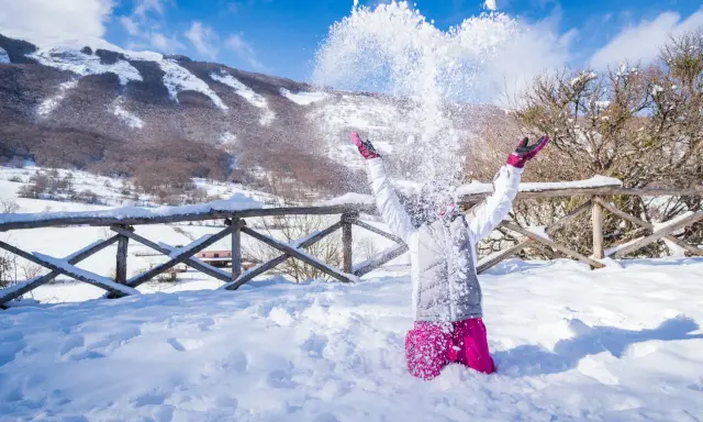 Bambina felice lancia neve in aria, con montagne innevate sullo sfondo. Perfetto per scoprire dove andare per la neve in Abruzzo.