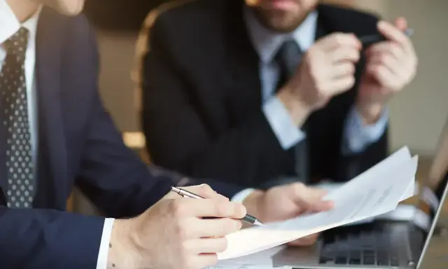 Dwóch mężczyzn w garniturach analizuje dokumenty. Jeden z nich wskazuje piórem na umowę, która może być naganą dla pracownika.