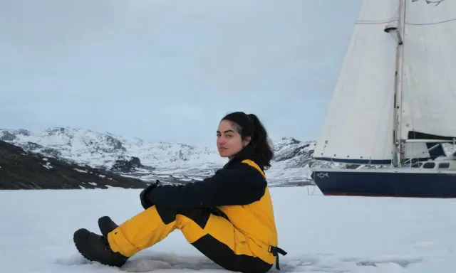 Tamara Klink, en tenue de ski jaune et noire, assise sur la neige, avec un voilier et des montagnes enneigées en arrière-plan.