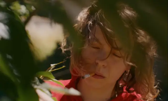 En närbild på Nina Widerberg med en cigarett i munnen, omgiven av gröna blad.