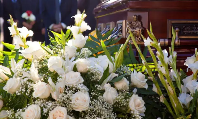 Elegancki wieniec pogrzebowy z białych róż i lilii, ozdobiony zielenią, przy trumnie. Cena 200 zł.