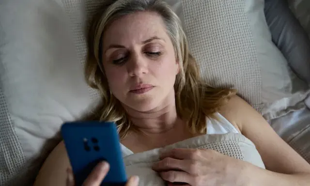 Kobieta leży w łóżku, trzymając telefon. Może to być związane z problemem oddawania moczu w nocy jeden raz.