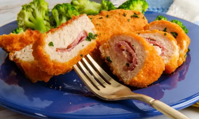 Kotlet po szwajcarsku, panierowany, z szynką w środku, podany z brokułami na niebieskim talerzu. Złoty widelec czeka na pierwszy kęs.
