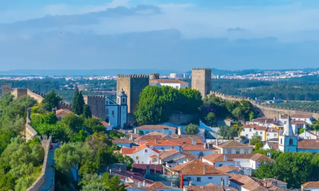 Malerische portugiesische Landschaft mit einer mittelalterlichen Burg und Stadtmauern, die sich über grüne Hügel erstrecken. Weiße Häuser mit Ziegeldächern und ein Kirchturm sind sichtbar.