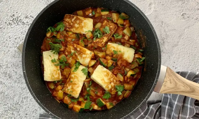 Smażony ser halloumi z warzywami w sosie pomidorowym, posypany natką pietruszki, w czarnej patelni.