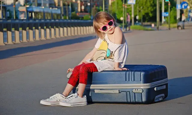 Dziewczynka w okularach siedzi na walizce, gotowa na wyjazd dziecka za granicę.