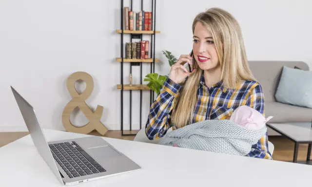 Kobieta z dzieckiem na rękach rozmawia przez telefon, pracując przy laptopie. To przykład pracy z orzeczeniem o niepełnosprawności, pokazujący elastyczność i możliwości.