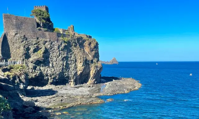Castello Normanno a Aci Trezza, un luogo da vedere. Roccioso promontorio sul mare blu, con un antico castello in pietra e vegetazione.