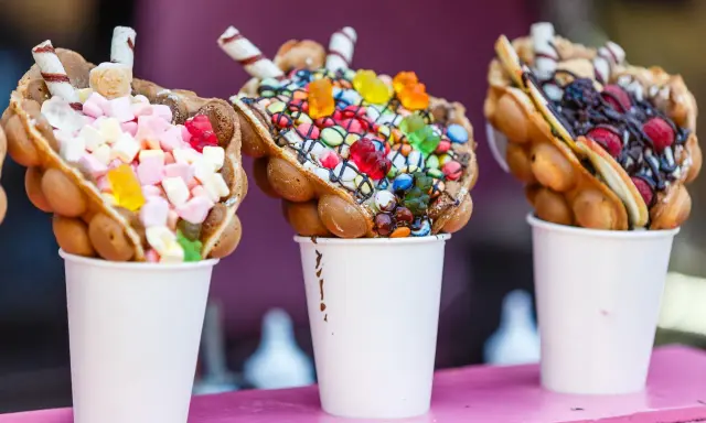 Trois bubble waffles colorés, garnis de bonbons, guimauves et fruits, présentés dans des gobelets blancs.