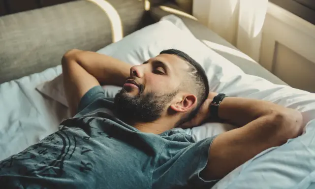 Mężczyzna z zarostem leży na łóżku z rękami za głową. Słońce wpada przez okno, ale jego oczy są zamknięte, jakby walczył z bezsennością.