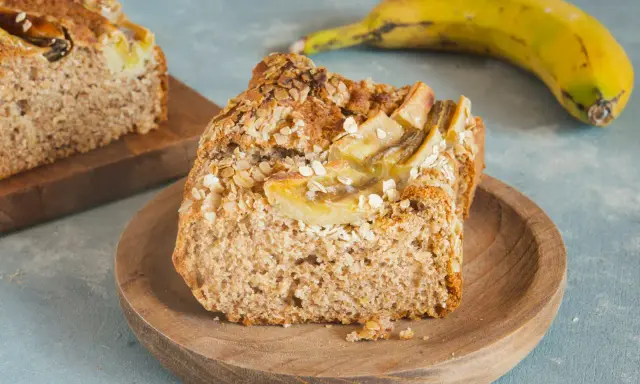 Pyszny chlebek bananowy fit z płatkami owsianymi na wierzchu, podany na drewnianym talerzyku. Obok leży dojrzały banan.