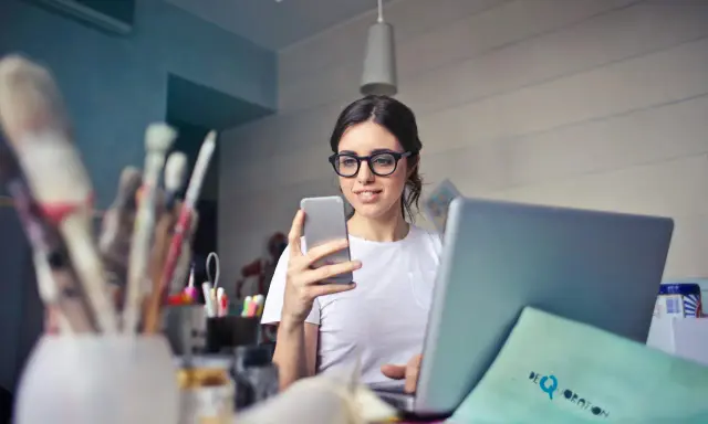 Kobieta w okularach do komputera przegląda telefon, siedząc przy laptopie.