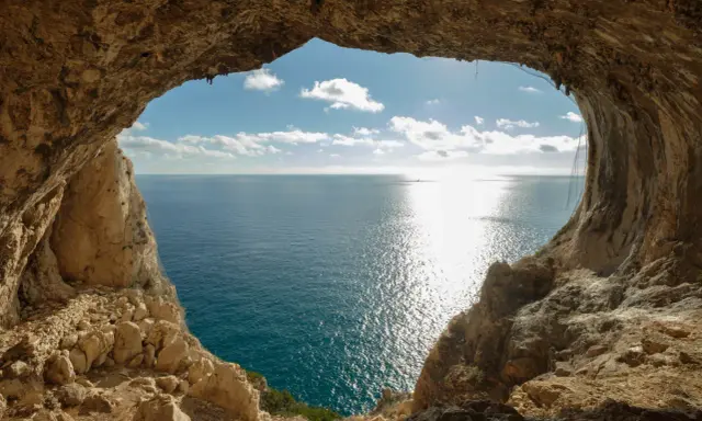 Vista dal mare da una grotta, con un sentiero del pellegrino che si intravede tra le rocce. Il sole brilla sull'acqua.