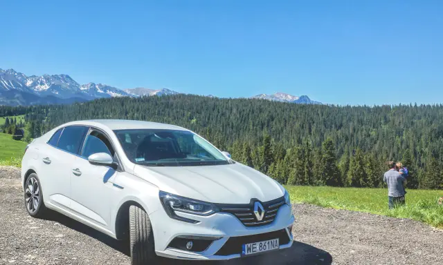 Biały Renault Megane Sedan zaparkowany na szutrowej drodze z widokiem na góry i las. Na trawie stoi mężczyzna z dzieckiem.