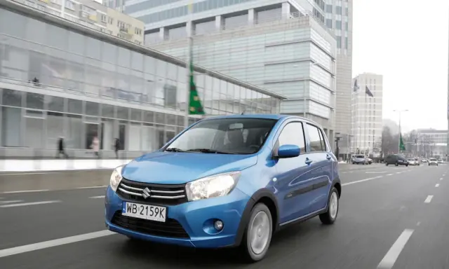 Niebieski Suzuki Celerio jedzie ulicą w mieście, mijając nowoczesne budynki i innych uczestników ruchu.