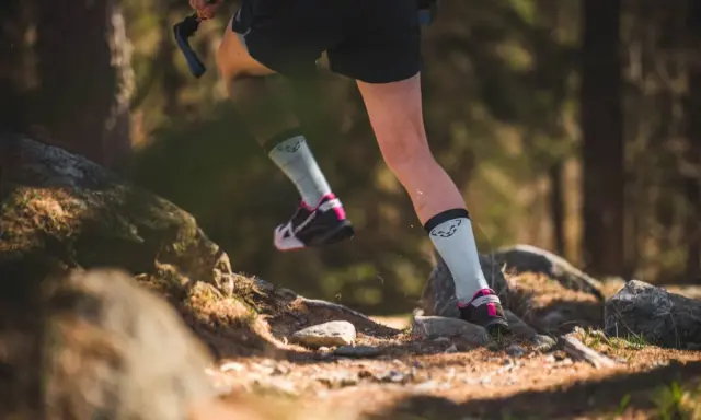 Beine einer Person beim **trail running** auf einem steinigen Waldweg. Leichte Sportschuhe und helle Socken mit Logo.