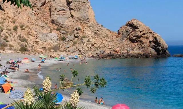 Playa de arena gris con gente disfrutando del sol y el mar, rodeada de acantilados. Un paraíso entre los pueblos de Granada con playa.