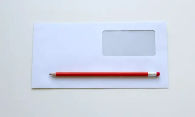 Biała koperta z okienkiem i czerwony ołówek. Proste narzędzia, by dowiedzieć się, jak wypełnić kopertę do wysłania.