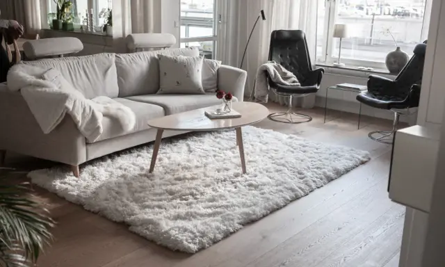 En fluffig, vit matta ligger framför en ljusgrå soffa och skapar en mysig känsla i vardagsrummet.