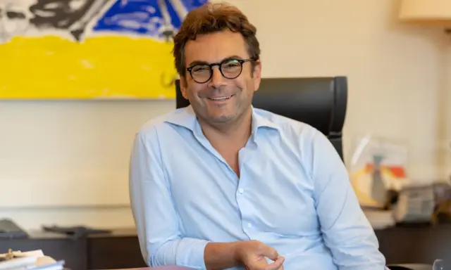 A man with glasses and a light blue shirt smiles while sitting at a desk with abstract art in the background.