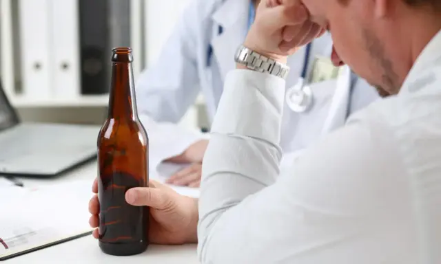 Mężczyzna trzyma butelkę piwa, siedząc przed lekarzem. Rozmowa sugeruje skierowanie na detoks alkoholowy.