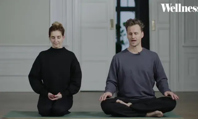 Två personer sitter på en yogamatta och mediterar. En kvinna i svart tröja och en man i grå tröja. Ta ett ögonblick att stanna upp.