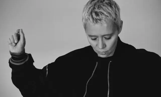 Jennifer Cardini, con i suoi capelli biondi corti e un'espressione pensierosa, alza un braccio con un gesto delicato. Indossa una giacca scura con zip.