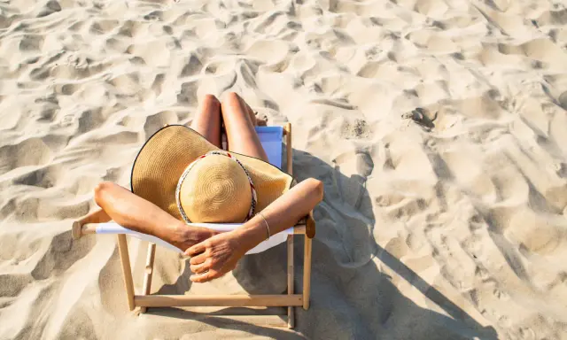 Kobieta w kapeluszu odpoczywa na leżaku na piaszczystej plaży. Idealne miejsce, by zapytać: gdzie na wakacje w lipcu?
