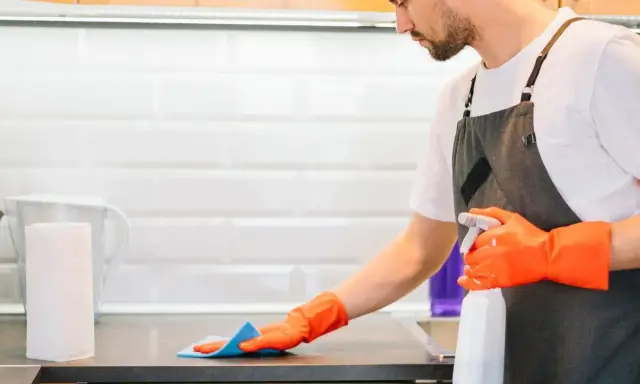 Hombre con guantes naranjas desinfecta la cocina, rociando la encimera con un spray limpiador.