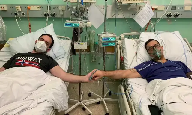 Dwóch mężczyzn w szpitalnych łóżkach, po operacji guza nerki, trzyma się za ręce. Jeden ma maskę tlenową, drugi zakrywa usta.