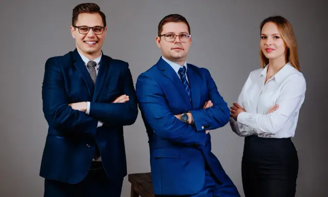 Trzy osoby w garniturach i białej koszuli, stojące na szarym tle, z rękami skrzyżowanymi na piersi.