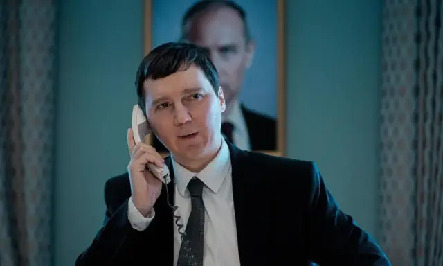 Un uomo in giacca e cravatta parla al telefono, con un ritratto di Putin sullo sfondo. Sembra il mago del Cremlino.