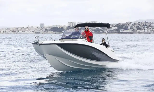 Bateau blanc et gris naviguant sur l'eau, avec trois personnes à bord. Le tableau de bord indique la puissance du moteur du bateau. Ville en arrière-plan.
