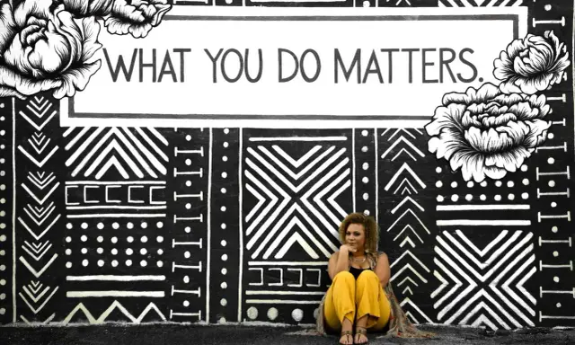 Kobieta siedzi przed muralem z napisem "WHAT YOU DO MATTERS". To inspirujący dialog motywujący.